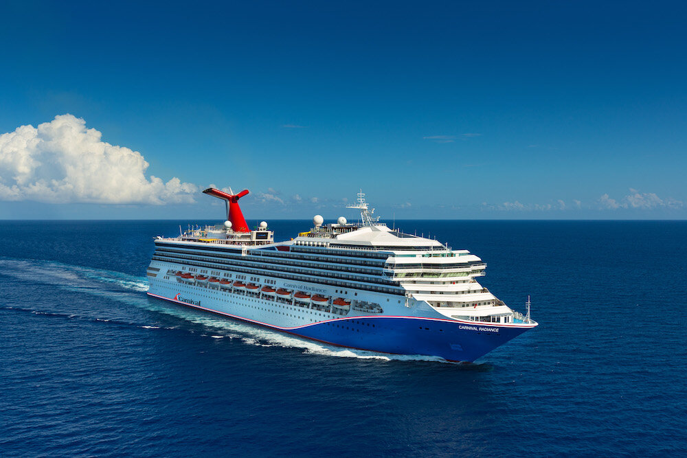 Carnival Radiance auf hoher See in der Seitenansicht mit charakteristischem roten Schornstein und blauem Rumpf.