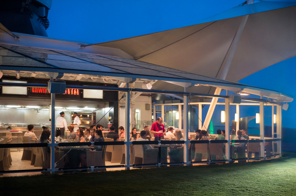 The Lawn Club Grill mit offener Küche und Gästen beim Speisen unter Sonnenschutze.