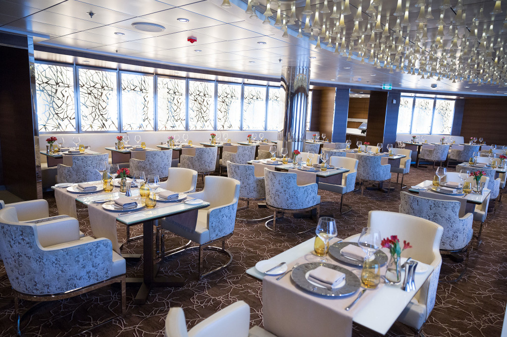 Moderner Speiseraum mit Fensterfront, eleganten blauen Sesseln und weiß gedeckten Tischen in der Luminae Restaurant des Schiffes.