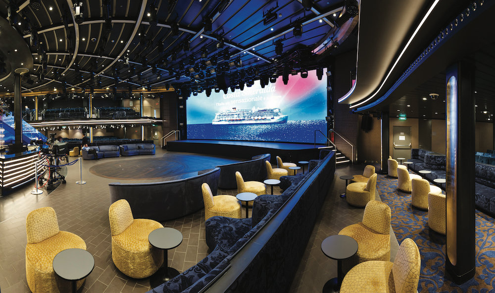 Entertainmentlounge auf einem Kreuzfahrtschiff mit goldenen Loungsesseln um eine runde Bühne und LED-Bildschirm