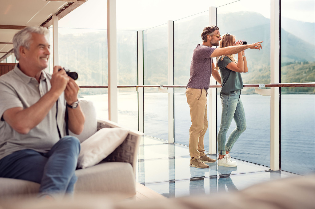 Gläserner Balkon mit Gästen beim Beobachten und Fotografieren einer sommerlichen Fjordlandschaft mit Bergen
