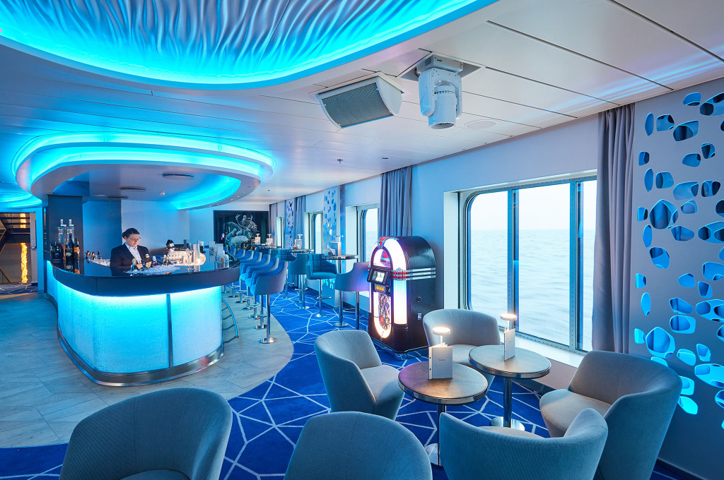 Bar mit blau beleuchtetem Wellendach, runder Theke, Jukebox und Sesseln vor Panoramafenster mit Meerblick