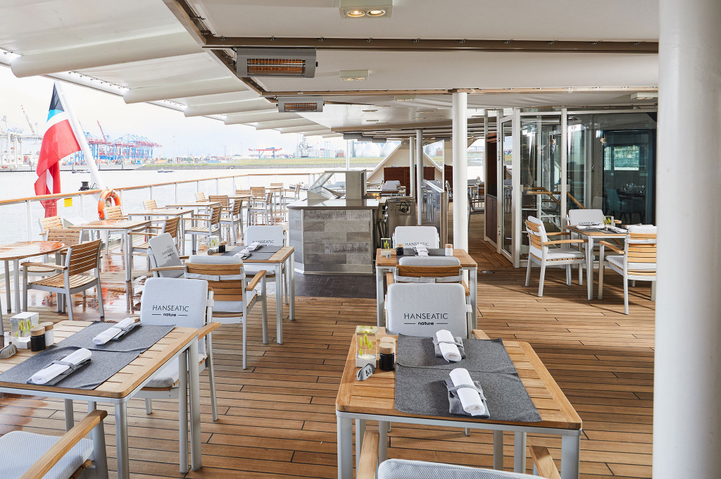 Outdoor-Terrasse des Lido-Restaurants mit Teakdeck, gedeckten Tischen und Blick auf Hafenkräne