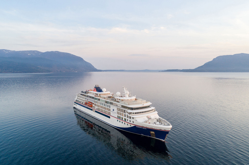 Luftaufnahme eines Expeditionskreuzfahrtschiffes mit weißem Rumpf und dunkelblauem Streifen in einem norwegischen Fjord