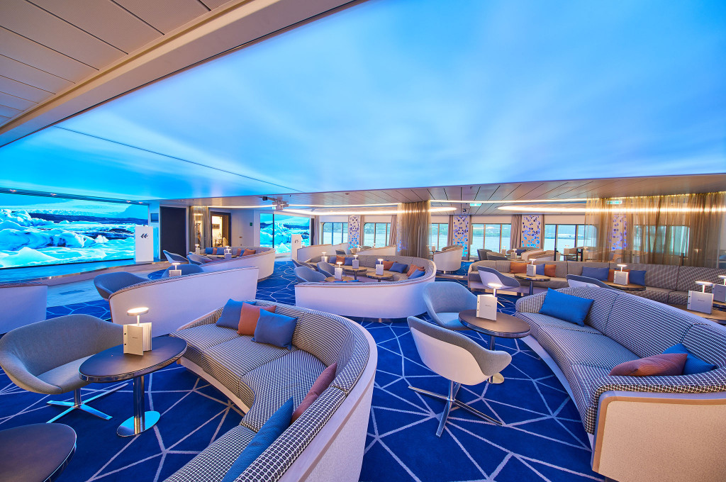 Lounge eines Expeditionsschiffes mit geschwungenen Sofas, Sesseln und Tischlampen vor einer beleuchteten Panoramawand mit Gletschermotiv