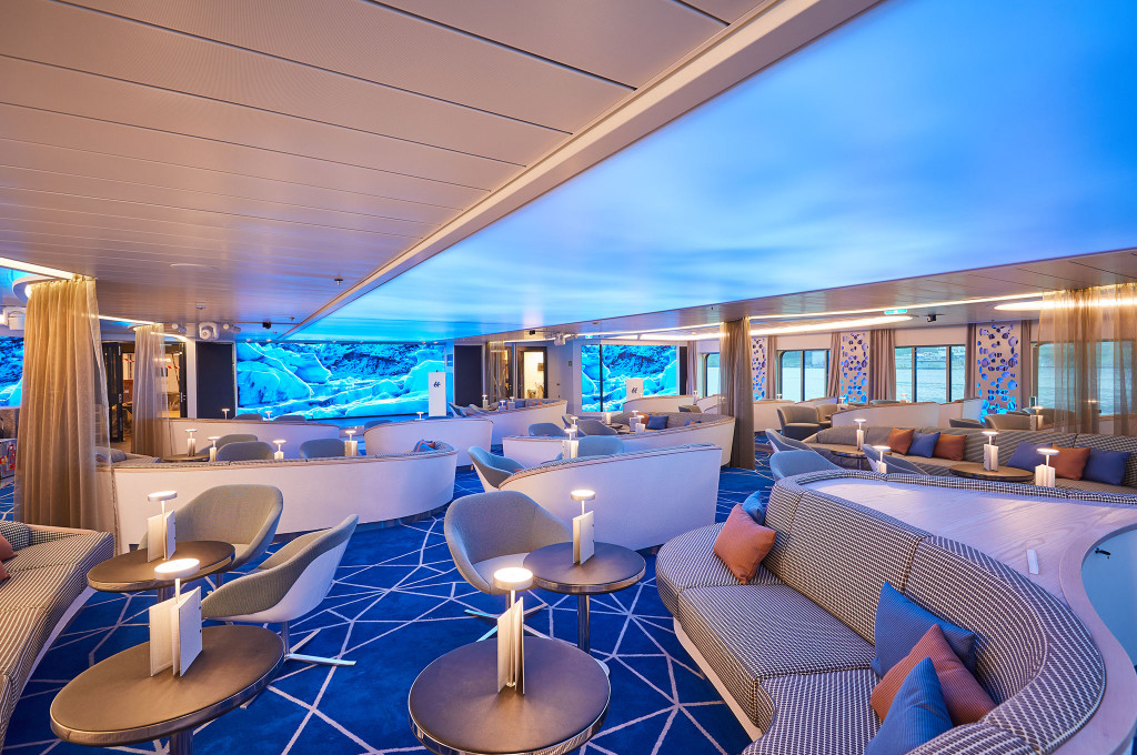 Großzügige Lounge eines Expeditionsschiffes mit blau beleuchtetem Deckenpaneel, gemusterten Rundsofas, Sesseln und Riesenleinwand mit arktischer Gletscherprojektion