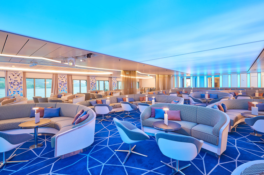Weitläufige Showlounge mit geschwungenen Sofas, Einzelsesseln, Tischlampen und Panoramafenstern mit Meerblick