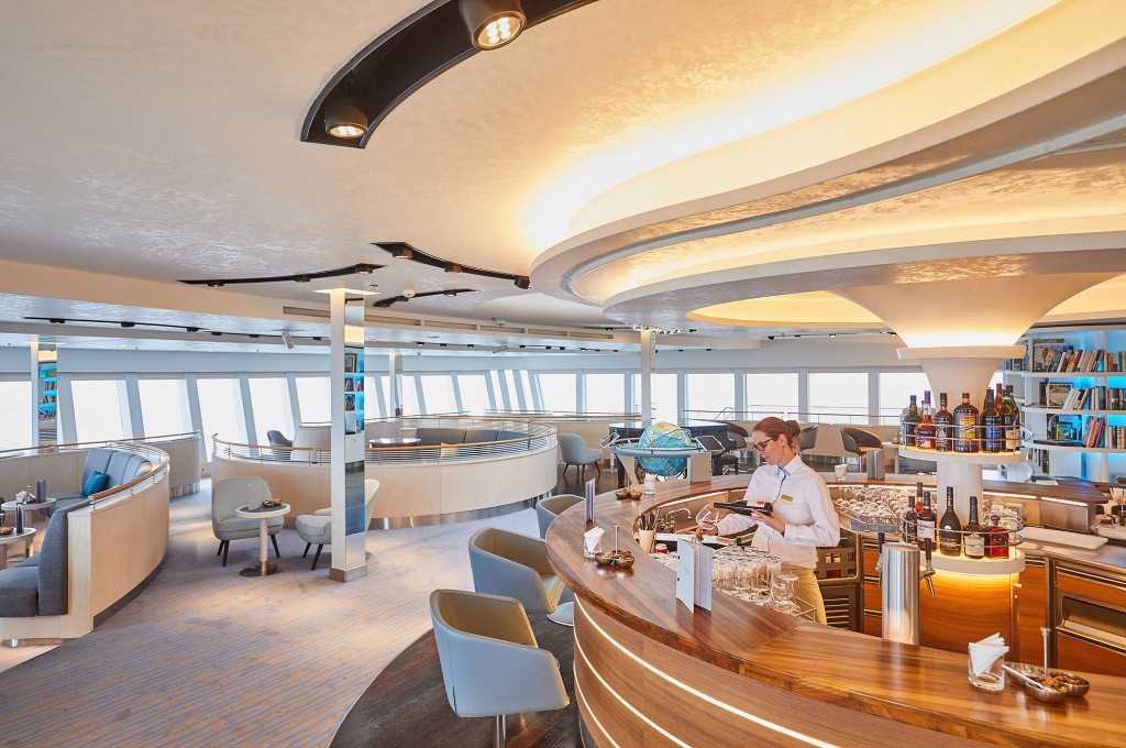 Observation Lounge mit geschwungener Holzbar, Barkeeperin, Globusdekor, Bücherregal und Panoramafenstern mit Meerblick