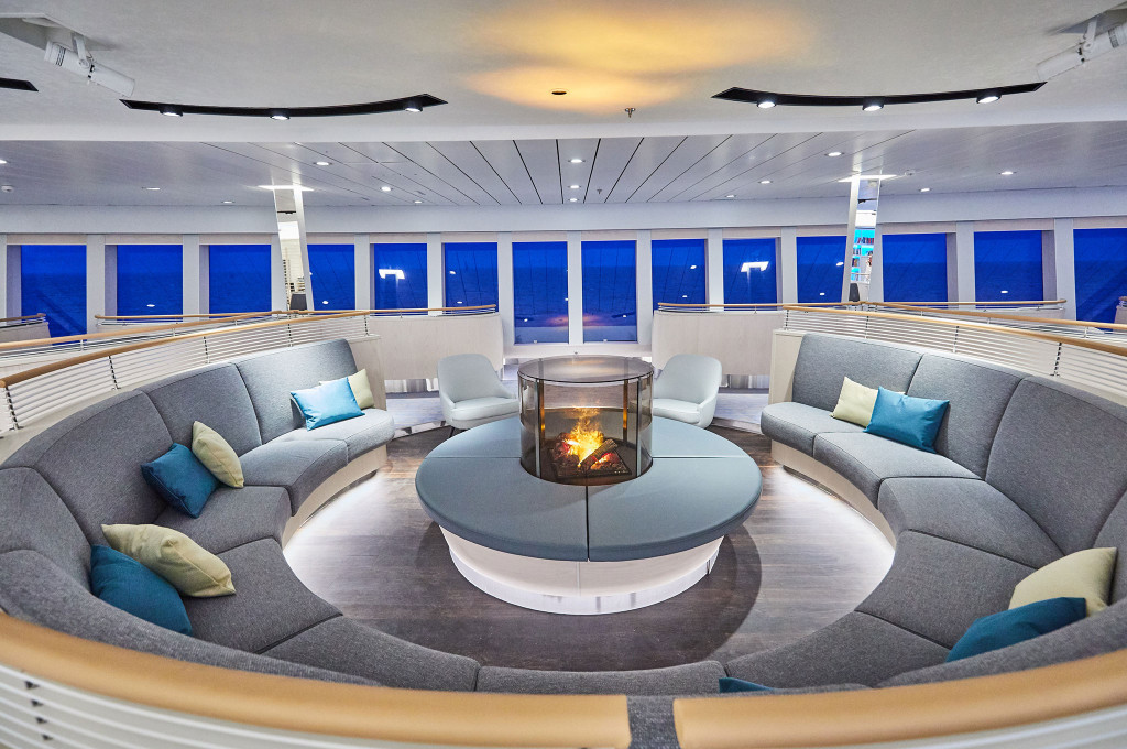 Kreisförmige Observation Lounge mit geschwungenen Sofas um einen zentralen Glaskamin und Panoramafenstern mit nächtlichem Meerblick