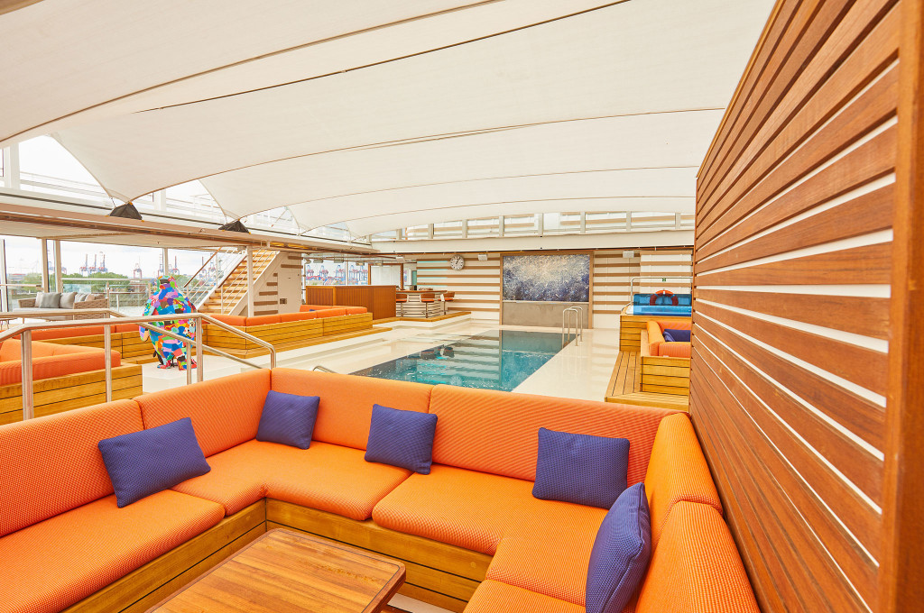 Überdachtes Pooldeck mit orangefarbenen Loungesofas, Pool, bunter Skulptur und Teakdeck im Hintergrund Hafenpanorama