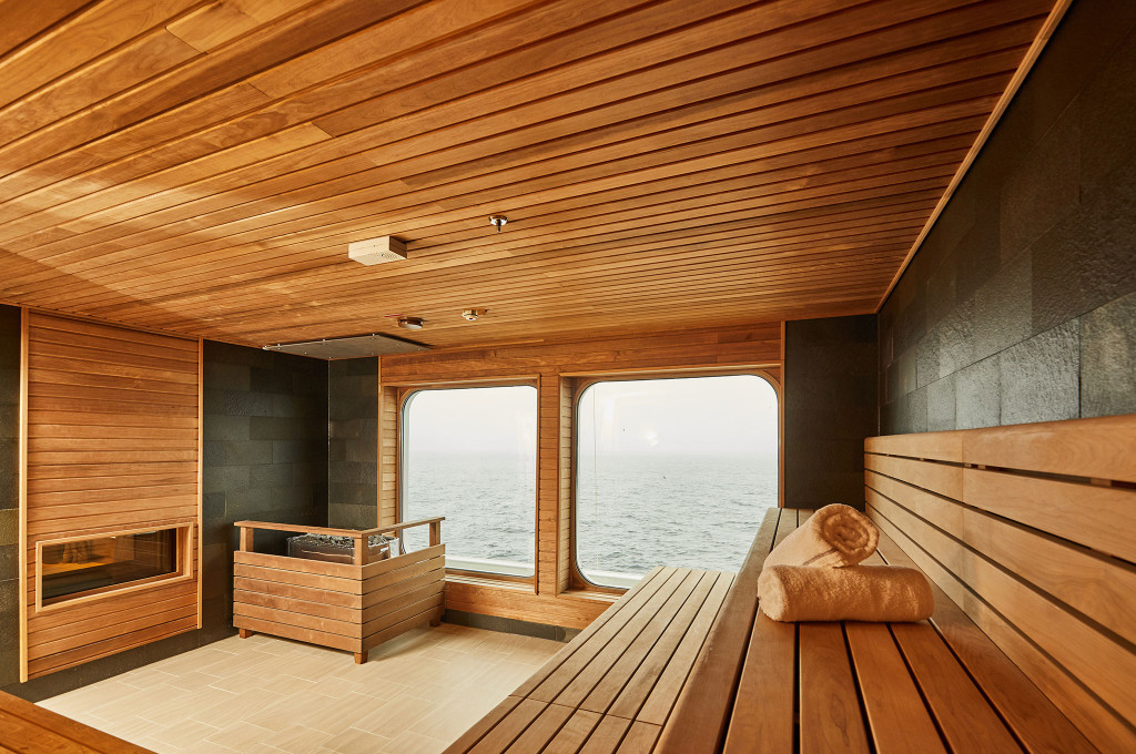 Holzsauna mit Zedernholzverkleidung, Saunasteinen, Holzbank mit Handtuch und Panoramafenstern mit Meerblick