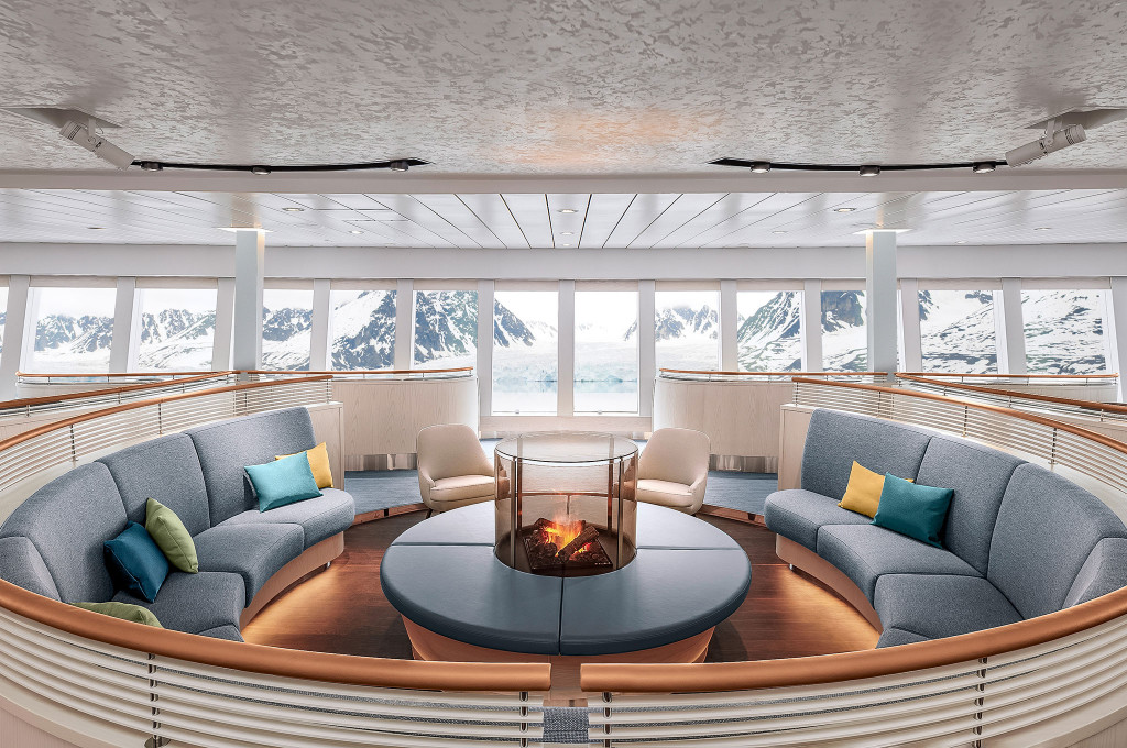 Kreisförmige Observation Lounge mit geschwungenen Sofas, zentralem Glaskamin und Panoramafenstern mit Blick auf arktische Schneeberge
