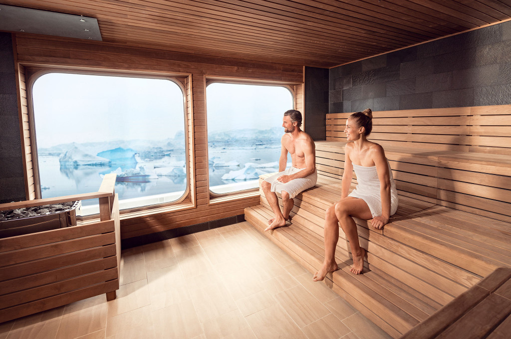 Holzsauna mit zwei Gästen in Handtüchern auf der Bank und Panoramafenstern mit Blick auf arktische Eisberge