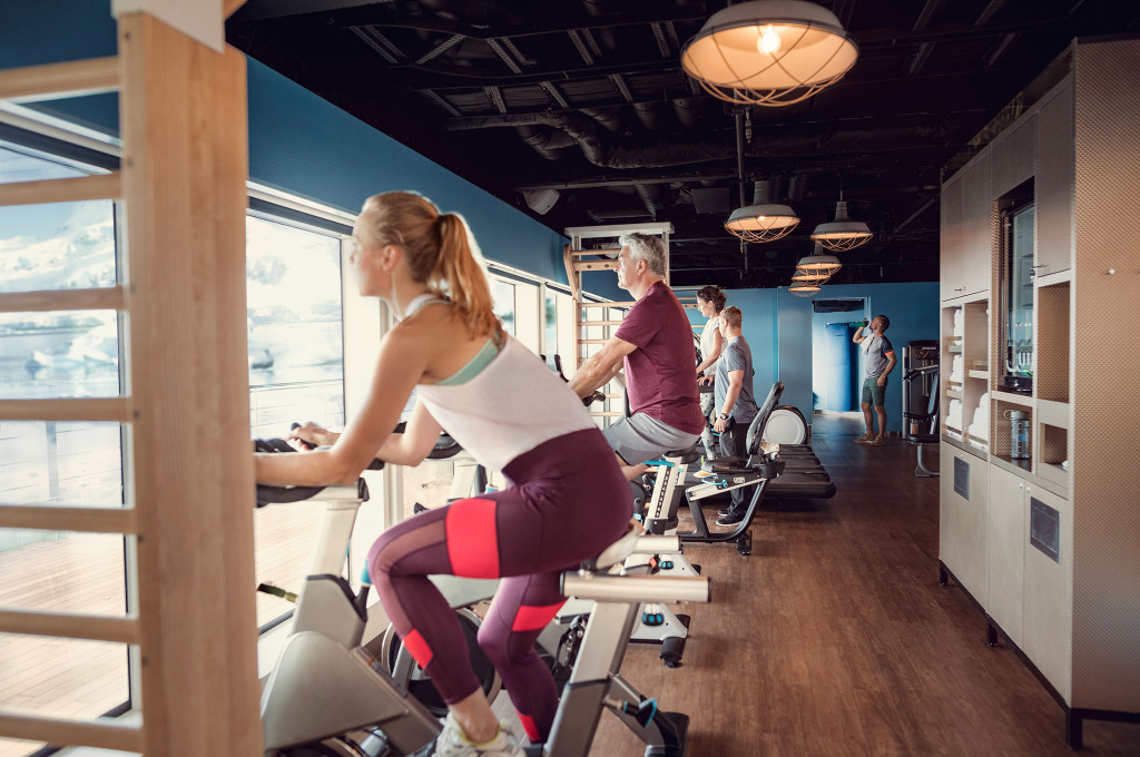 Fitnessraum mit Gästen auf Spinningbikes und Ergometern vor Panoramafenstern mit arktischer Eislandschaft