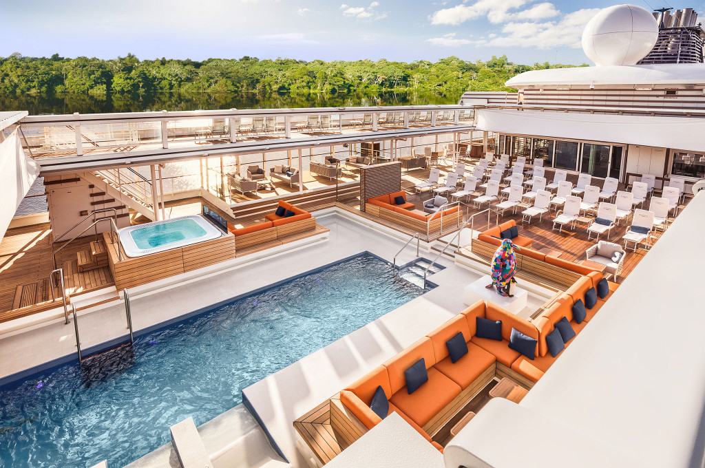 Weitläufiges Pooldeck mit Pool, Whirlpool, orangefarbenen Loungesofas, Liegestühlen und bunter Skulptur vor tropischer Amazonas-Flusslandschaft
