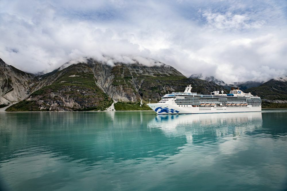 Kreuzfahrtschiff in einem türkisfarbenen Fjord vor wolkenverhangenen Bergen mit Spiegelung im Wasser