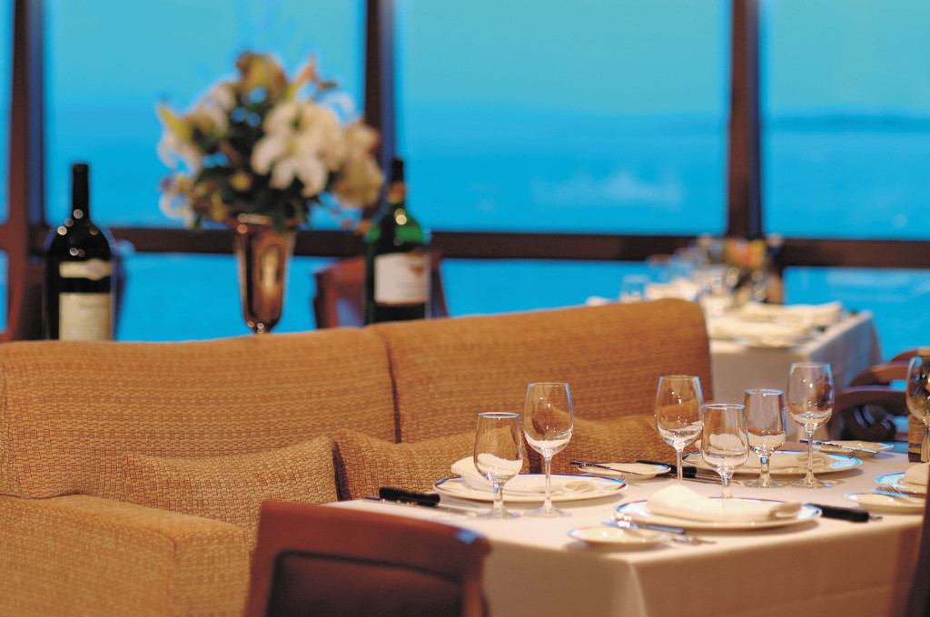Eingedeckter Tisch im Spezialitätenrestaurant mit Weinflaschen und Blumenschmuck vor Panoramafenstern mit Meerblick.
