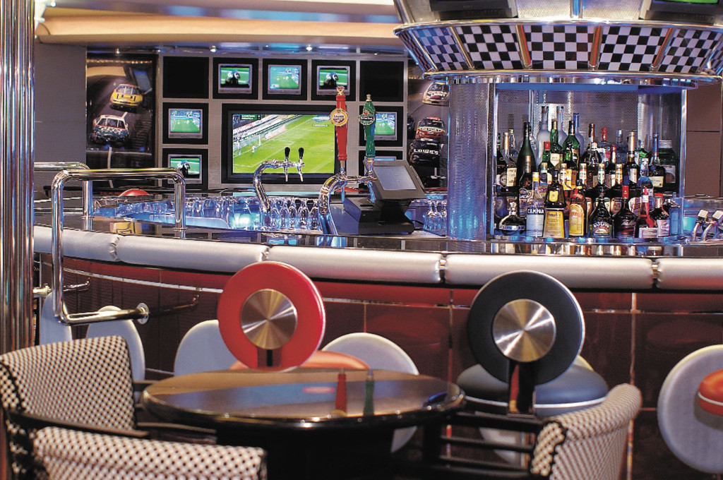 Sportbar mit Zapfhähnen, Flaschenregal, mehreren Fernsehern und bunten Barhockern an Bord des Schiffes.