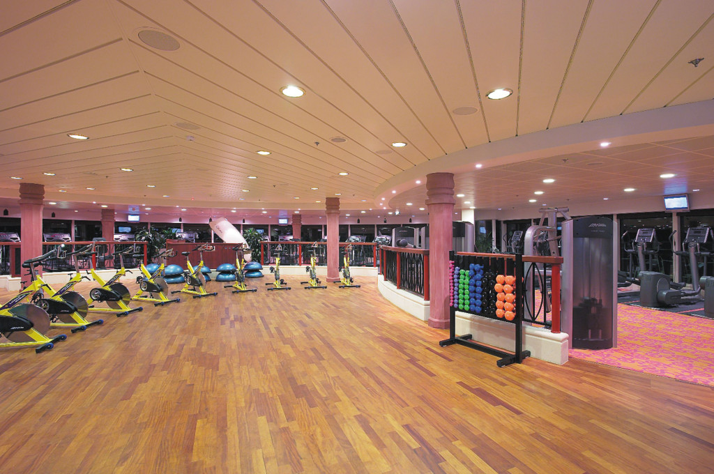 Fitnesscenter mit Reihen gelber Ergometer, Hantelständer und Cardiogeräten auf Holzboden an Bord.