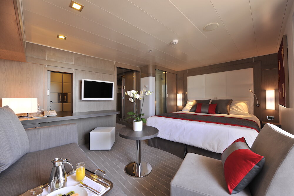 Elegant eingerichtete Suite auf einem Kreuzfahrtschiff mit Doppelbett, Sitzbereich und Frühstückstablett