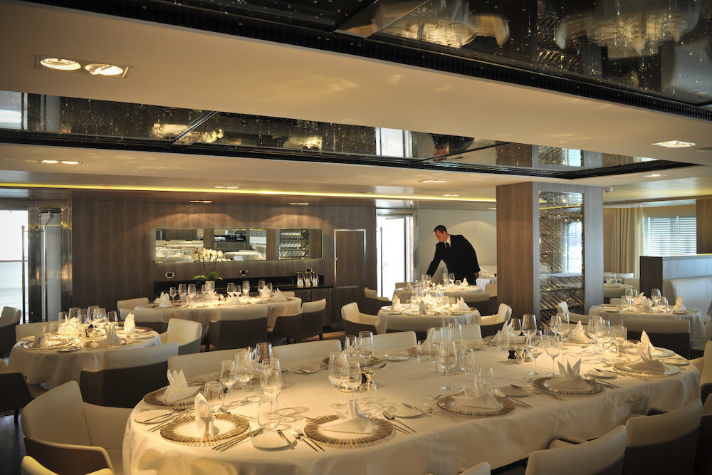 Elegantes Spezialitätenrestaurant mit weiß gedeckten Tischen, Spiegeldecke, Weinregal und Kellner beim Eindecken