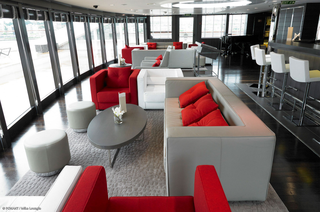 Panorama Lounge mit roten und weißen Sitzmöbeln, angrenzender Bar mit Barhockern und bodentiefen Fenstern.