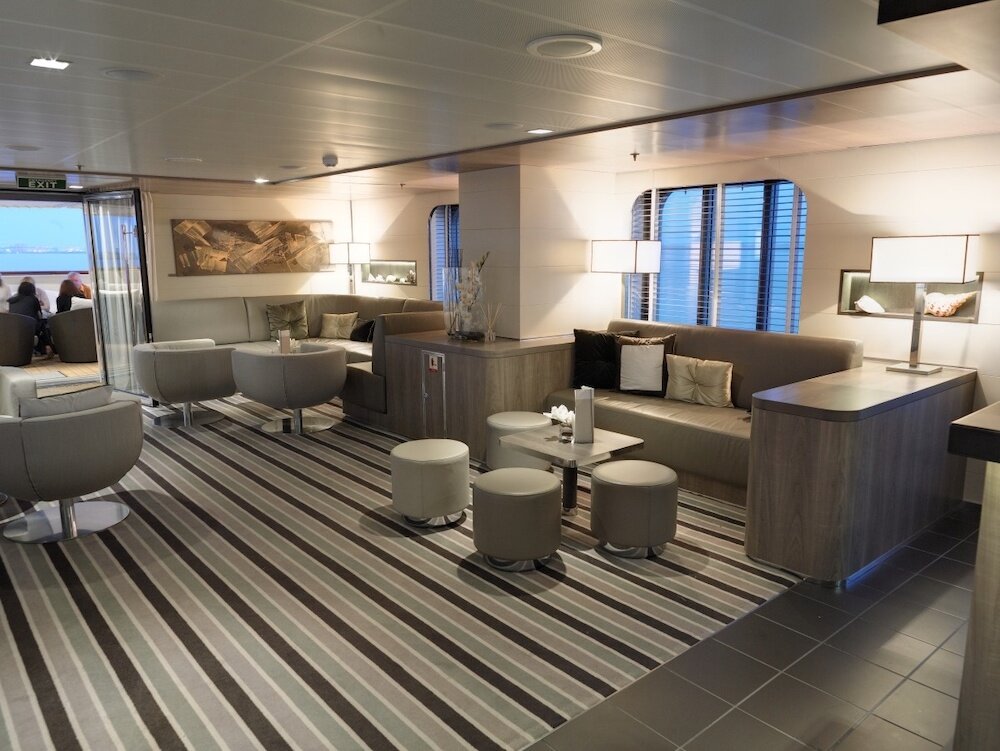Moderne Lounge mit beigen Sofas, runden Hockern, Streifenteppich und Panoramafenstern mit Meerblick auf einem Hochseekreuzfahrtschiff