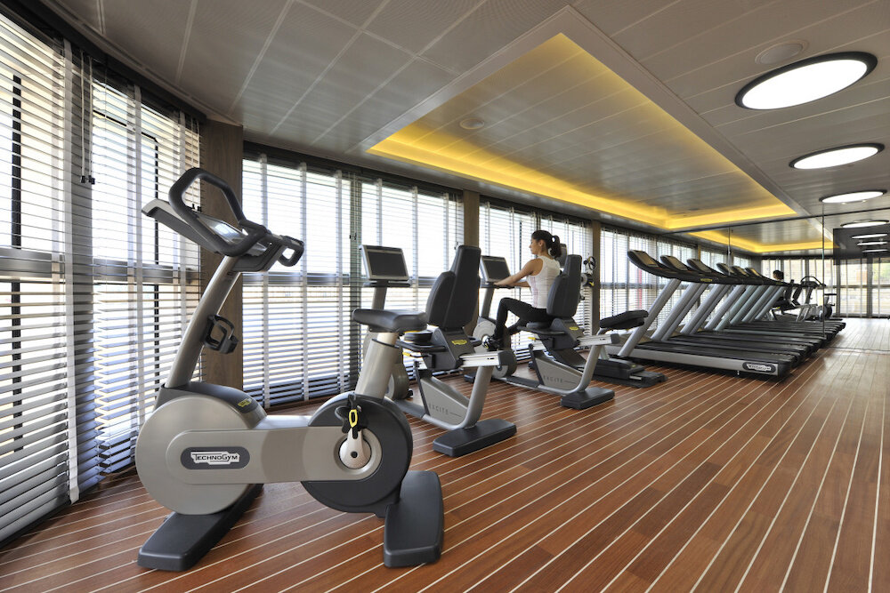 Moderner Fitnessraum mit Technogym-Fahrrad- und Liegeergometern, Laufbändern, Holzboden und großen Fensterfronten mit Jalousien
