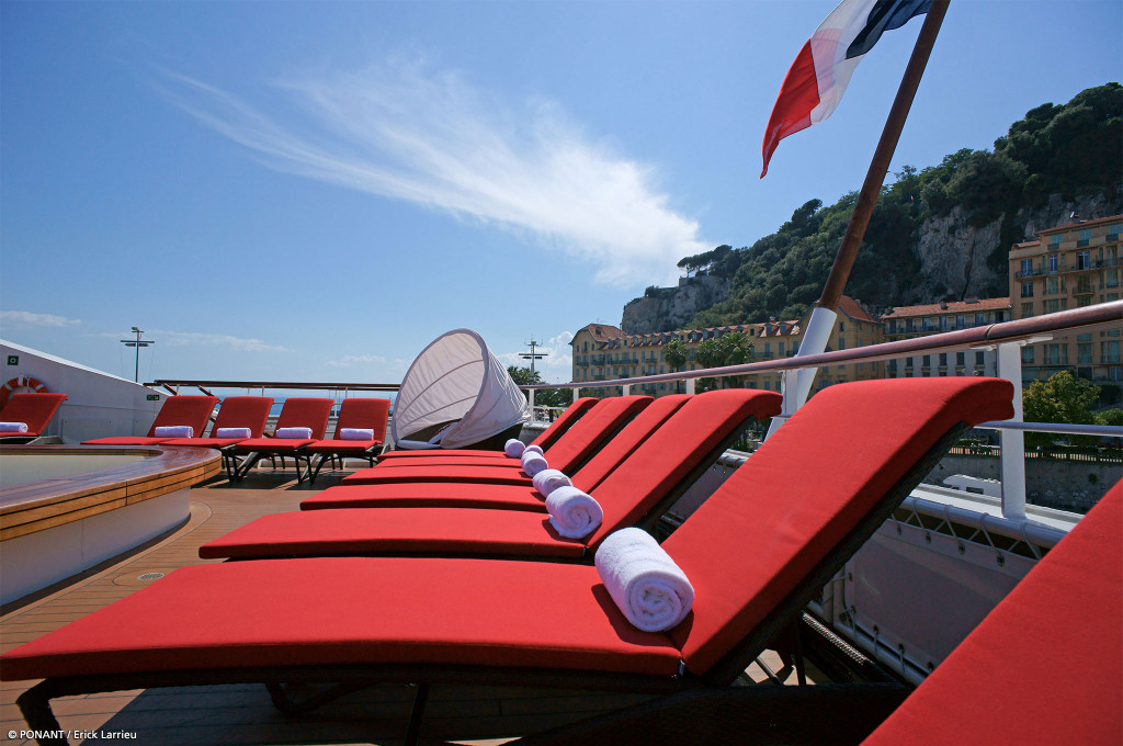 Sonnendeck mit roten Liegestühlen und Whirlpool, französische Flagge weht vor Hafenstadt mit Bergen.