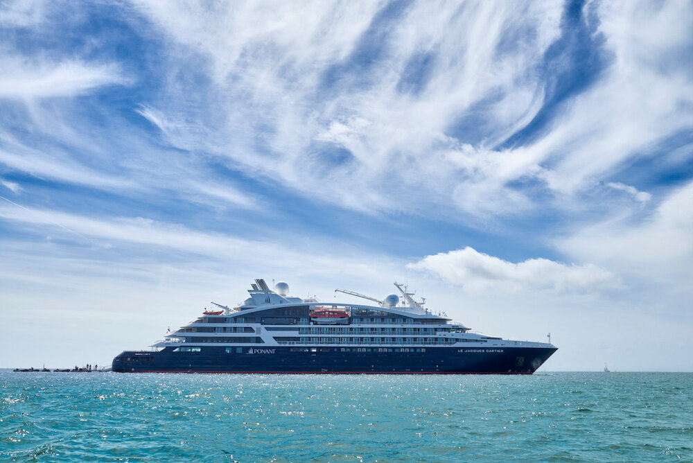 Modernes Luxus-Expeditionsschiff mit dunklem Rumpf ankert auf türkisblauem Meer unter dramatischem Wolkenhimmel