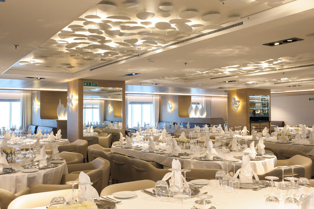 Helles Hauptrestaurant mit runden Tischen, beigen Lederstühlen, skulpturaler Deckeninstallation und Wandleuchten