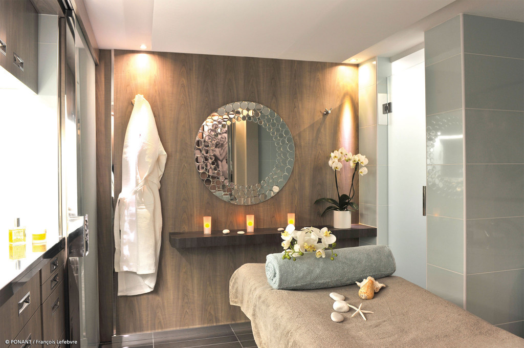 Spa-Behandlungsraum mit Massageliege, rundem Spiegel und Bademantel an Bord eines Kreuzfahrtschiffs.