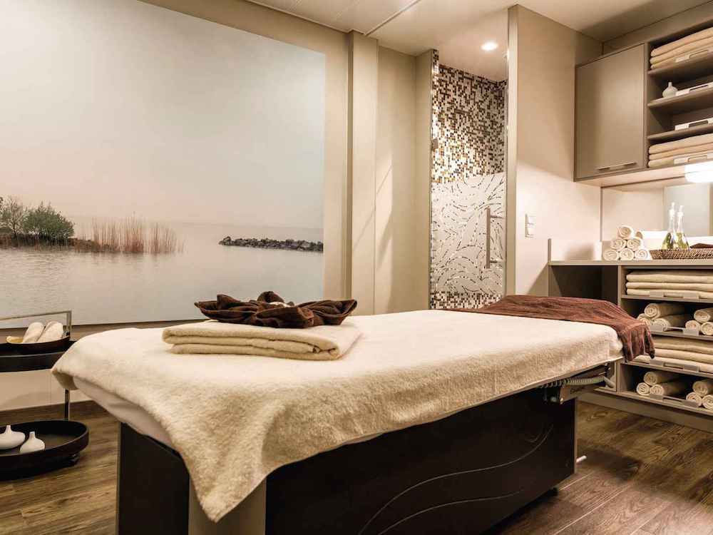 Spa-Behandlungsraum der Mein Schiff 3 mit Massageliege, Mosaikdetails, Landschafts-Wandbild und Handtuchregal