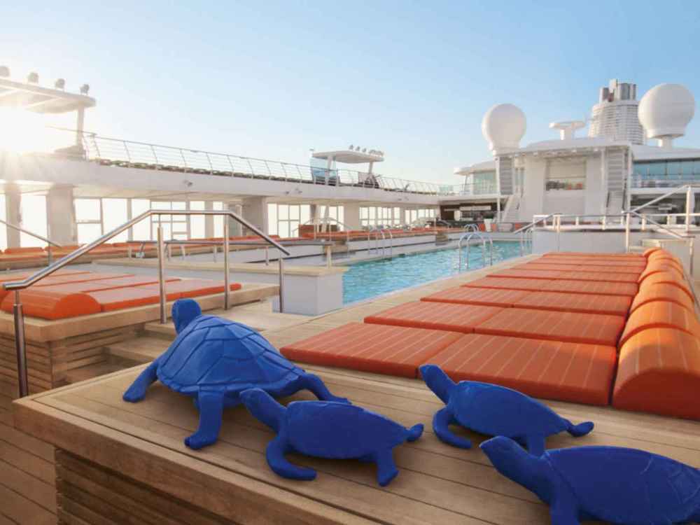 Pooldeck eines Kreuzfahrtschiffes mit Pool, orangefarbenen Liegestühlen und dekorativen Schildkröten-Kissen im Abendlicht
