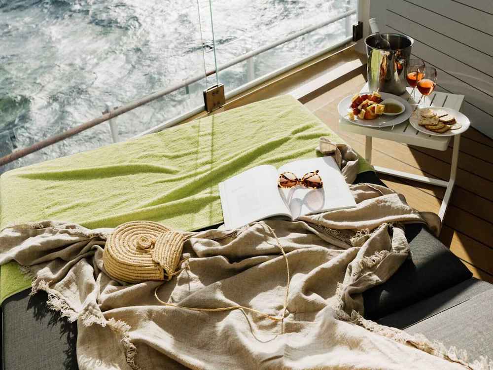 Kabinenbalkon mit Liegestuhl, Leinendecke, Buch und einem Beistelltisch mit Champagnerkühler und Snacktellern vor dem Meer