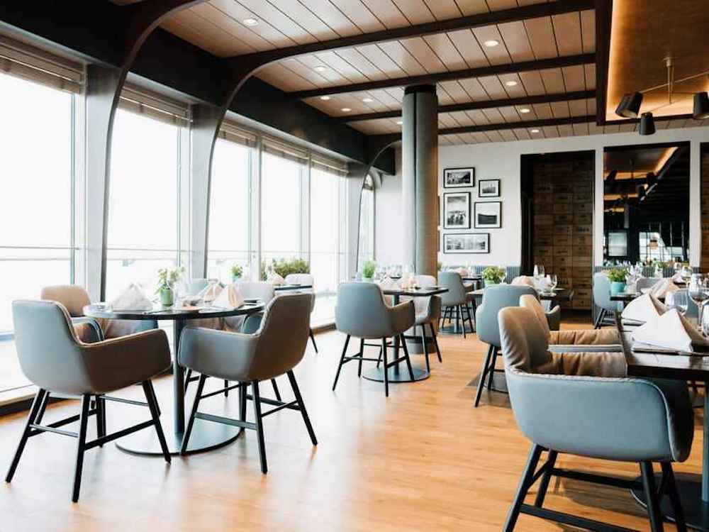 Helles modernes Restaurant mit hellgrauen Sesseln, dunklen Rundtischen und großen Panoramafenstern mit Meerblick auf einem Hochseekreuzfahrtschiff