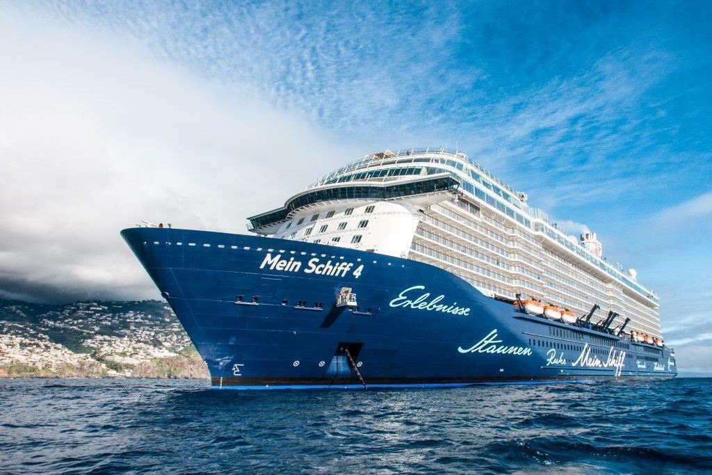 Kreuzfahrtschiff in voller Fahrt vor einer bergigen Küste bei bewölktem Himmel