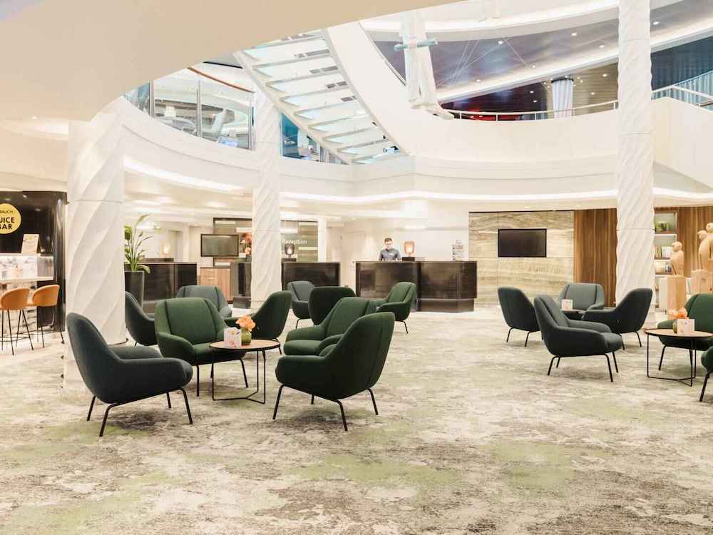 Mehrstöckiges Atrium mit geschwungenen weißen Säulen, grünen Sesseln, Rezeptionstresen und Glasgeländer auf einem Hochseekreuzfahrtschiff