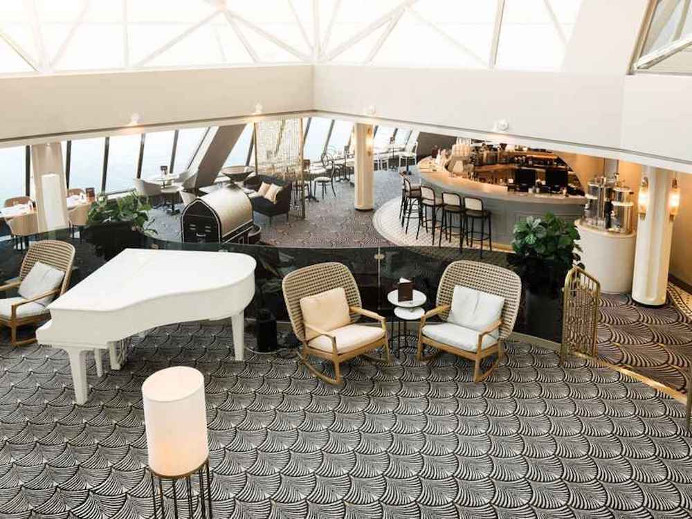 Weitläufige Lounge mit weißem Konzertflügel, Rattansesseln, geschwungenem Bartresen und großen Panoramafenstern mit Meerblick auf einem Kreuzfahrtschiff