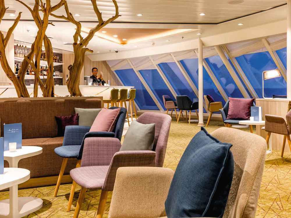 Moderne Lounge mit bunten Sessel, Treibholzinstallation und großen Schrägfenstern auf einem TUI-Kreuzfahrtschiff