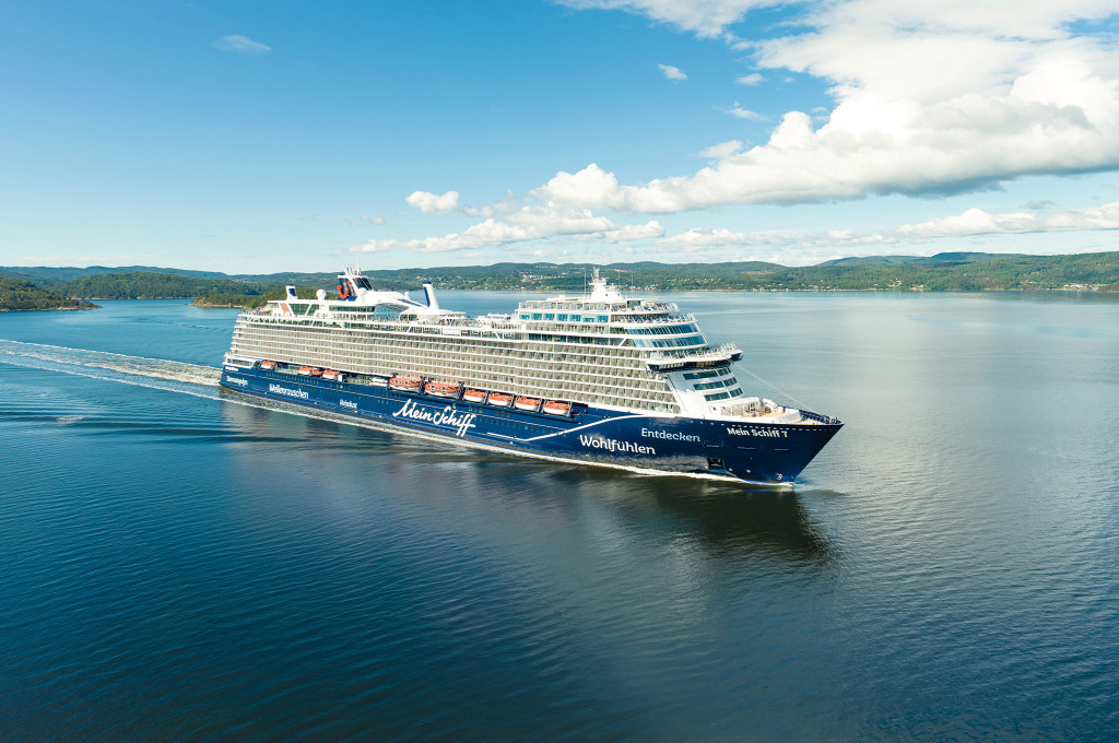 Kreuzfahrtschiff Mein Schiff 7 auf ruhigem Wasser vor einer bewaldeten Hügellandschaft.