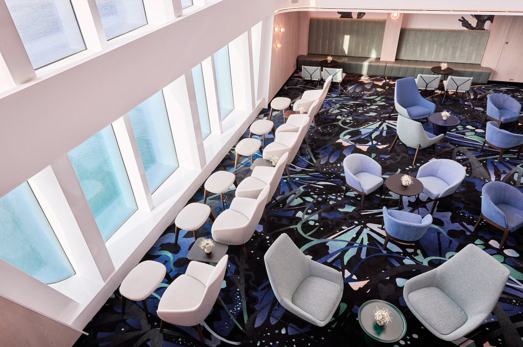 Blick von der Galerie in eine Lounge mit gepolsterten Sesseln, kleinen Tischen und großen Seitenfenstern.