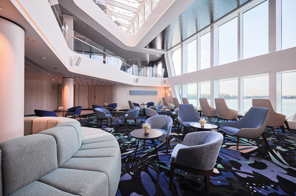Helle Lounge mit blauen und grauen Sesseln, runden Tischen und bodentiefen Fenstern mit Blick auf das Wasser.