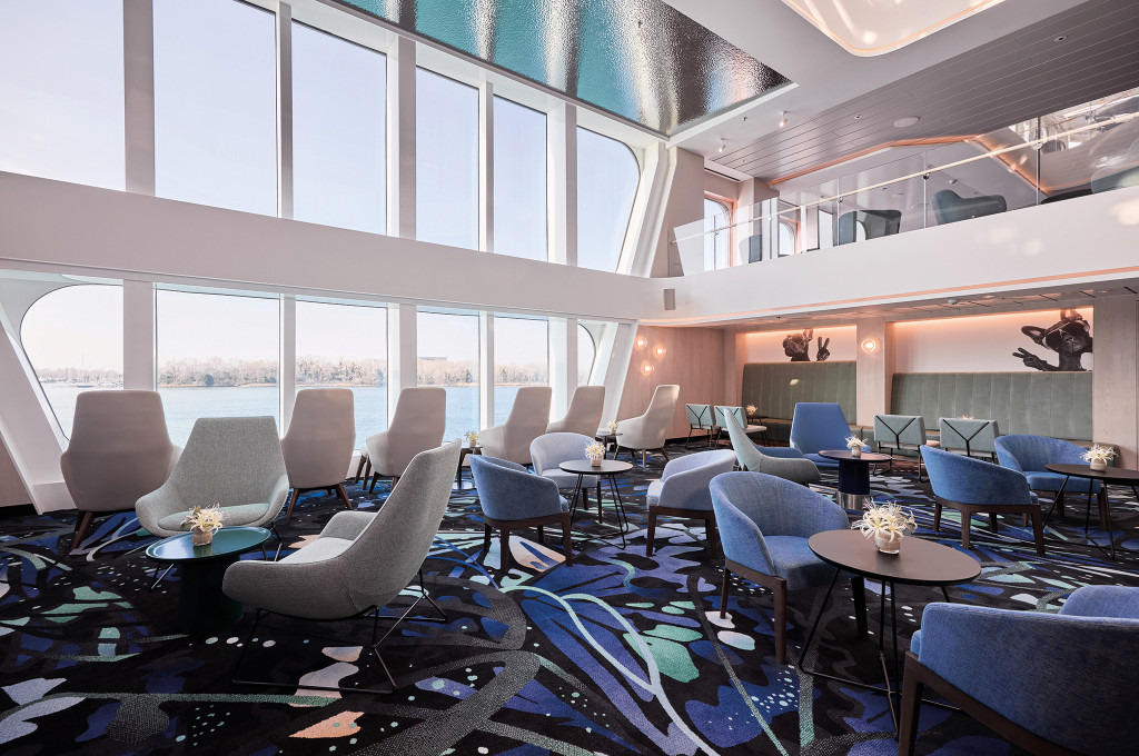 Doppelgeschossige Lounge mit grauen und blauen Sesseln, runden Tischen und raumhohen Fenstern mit Flussblick.