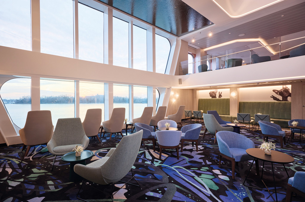 Mehrstöckiger Lounge- und Atriumbereich mit Sesseln, runden Tischen und bodentiefen Panoramafenstern mit Blick auf den Fluss.