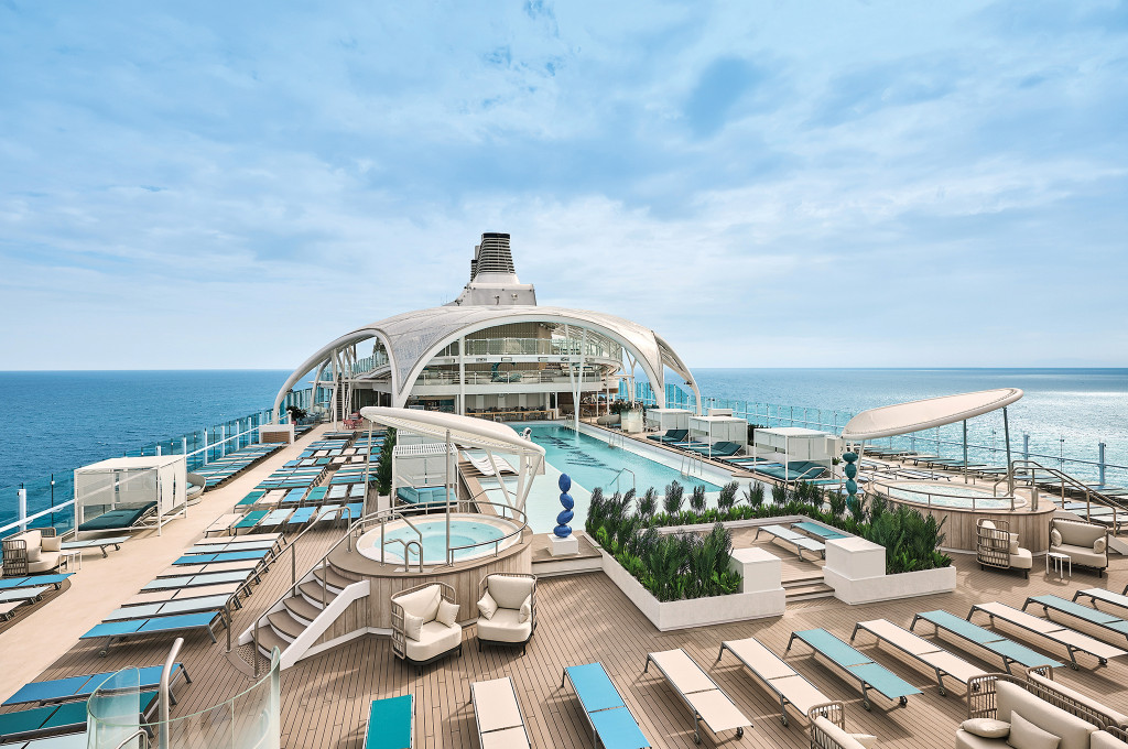 Pooldeck mit Außenpool, Whirlpool, Loungemöbeln und zahlreichen Liegestühlen unter Glasdachkonstruktion mit Blick auf das Meer.