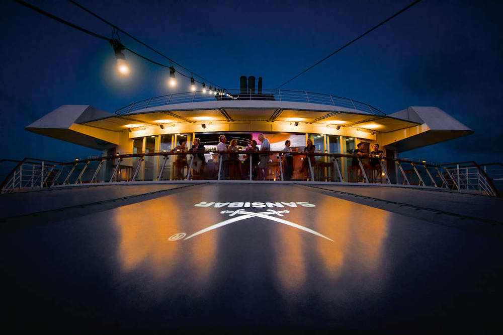 Sansibar-Bar auf dem Außendeck der MS Europa am Abend mit beleuchteten Gästen, Lichterketten und Sansibar-Logo-Projektion auf dem Deck
