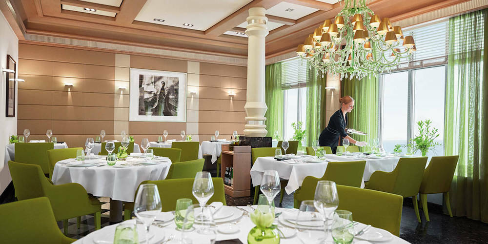 Restaurant Serenissima auf der MS Europa 2 mit grünen Stühlen, gedeckten Tischen, Kronleuchter und Panoramafenstern.