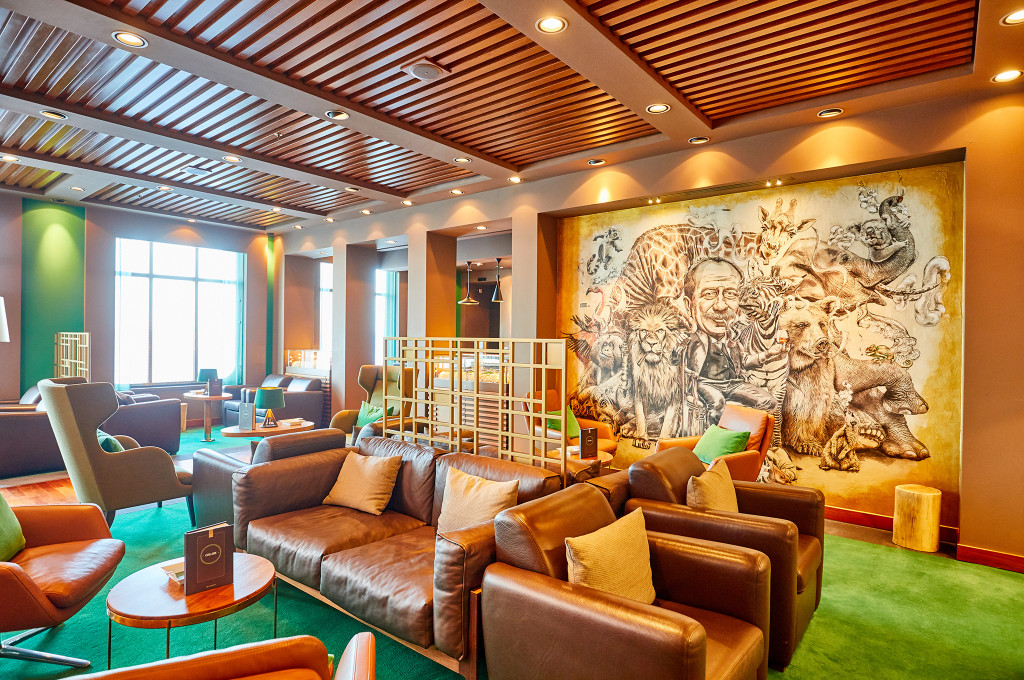 Weitläufige Collins-Lounge mit braunen Ledersofas, großem Tier-Wandgemälde, grünem Teppich und warmer Holzdecke