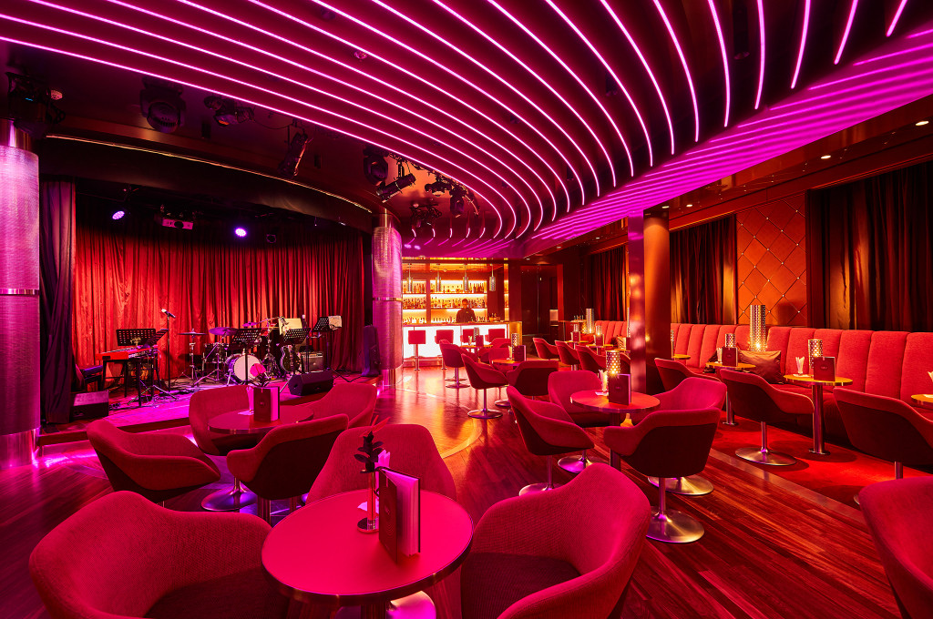 Nightclub mit pinker Neondeckenbeleuchtung, roten Sesseln, Bühne mit Schlagzeug und Roten Vorhängen sowie beleuchteter Bar